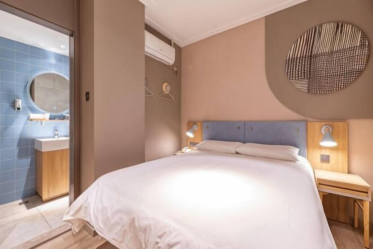 A bedroom with a bed, bedside table, wall-mounted lamp, and a bathroom with a sink and mirror.