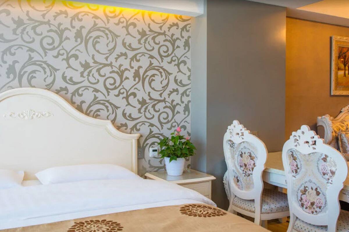 A hotel room with a bed, a nightstand with a potted plant, and two ornate chairs.
