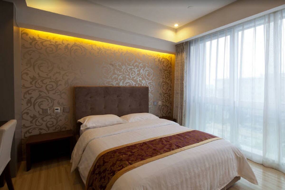 A hotel room with a large bed, a bench, and a view of the city through sheer curtains.