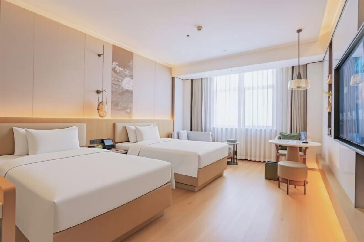 A hotel room with two beds, a small table, a chair, and a large window with curtains.