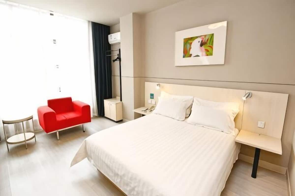 A hotel room with a large bed, a red chair, a bedside table, a nightstand, and a wall-mounted air conditioning unit.