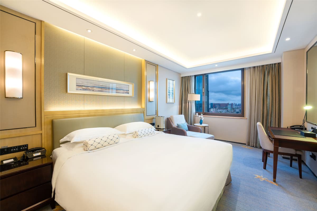 Une chambre d’hôtel avec un grand lit, un bureau, une chaise et une vue sur la ville par la fenêtre.