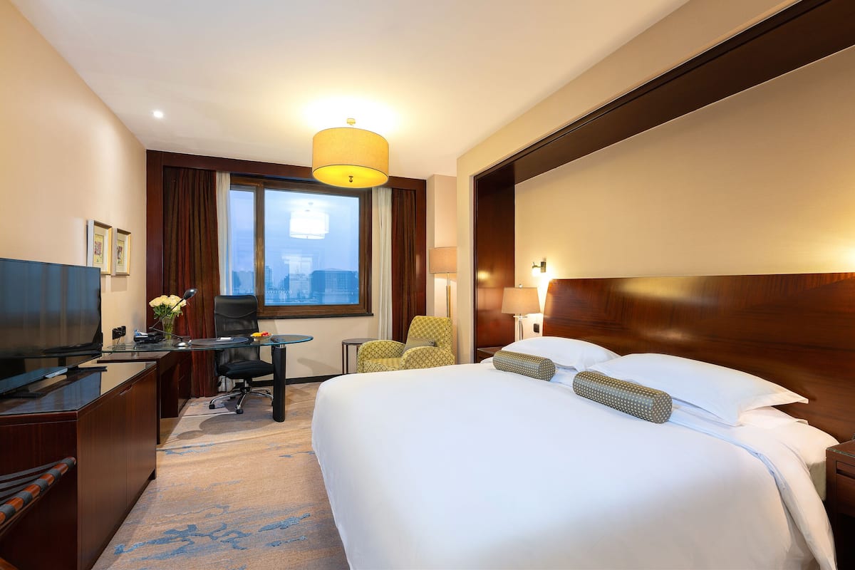 Une chambre d’hôtel avec un grand lit, un bureau doté d’un téléviseur à écran plat, une chaise, une lampe et une vue sur la ville par la fenêtre.
