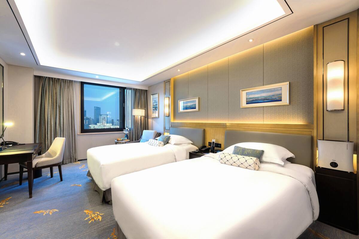 Une chambre d’hôtel avec deux lits, un bureau, une chaise, une lampe et une vue sur la ville.