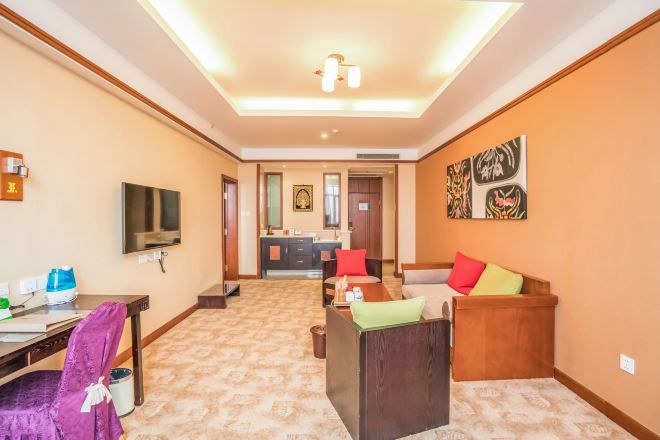 A hotel room with a television, a desk with a chair, a sofa with cushions, and a hallway leading to a bathroom and kitchen area.