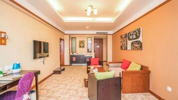 A hotel room with a television, a desk with a chair, a sofa with cushions, and a hallway leading to a bathroom and kitchen area.