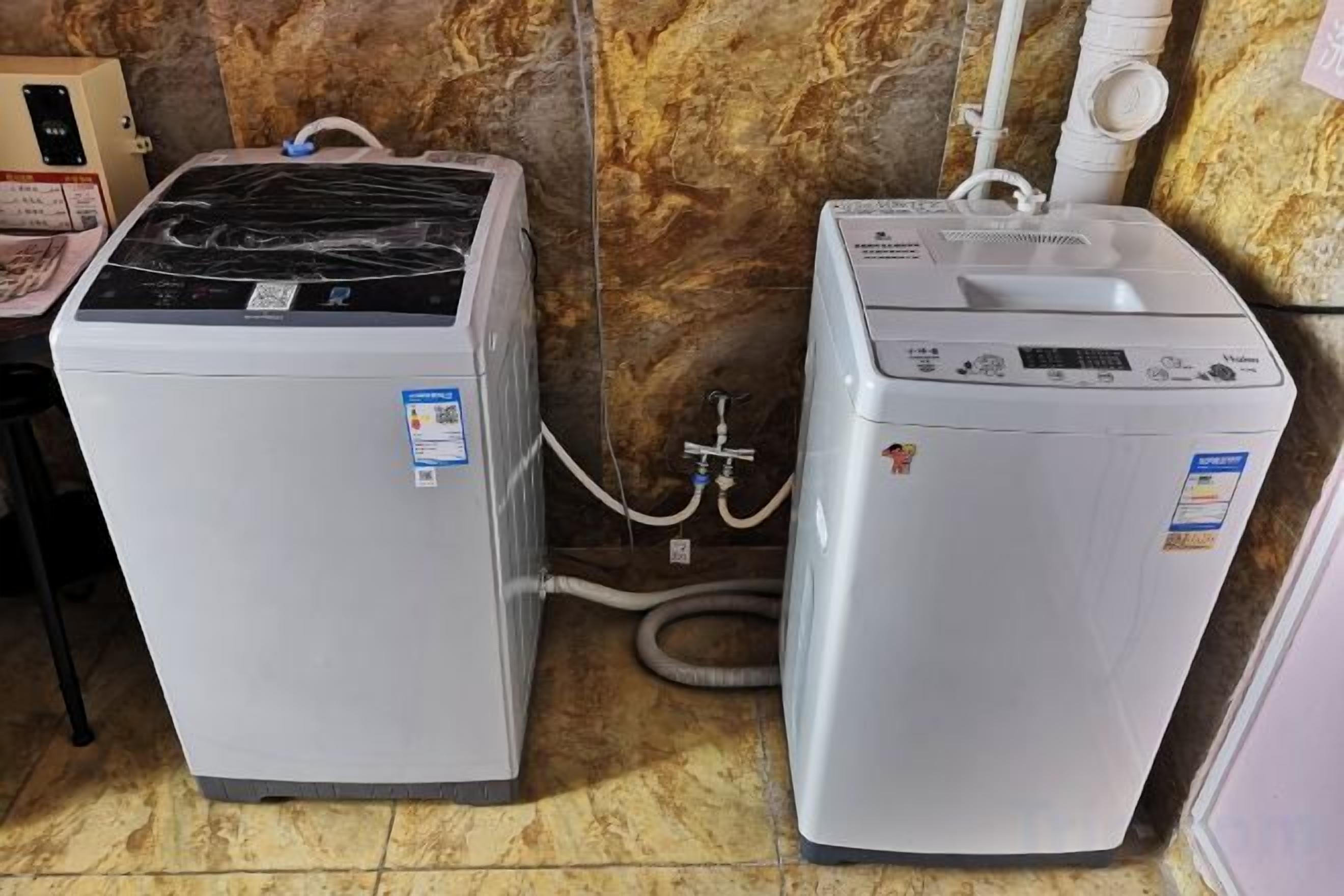Two white appliances, likely a washer and dryer, in a corner with a marble wall and a tiled floor.