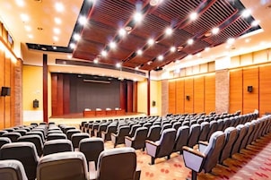 Un salón de conferencias con filas de sillas, un escenario y una pantalla grande.