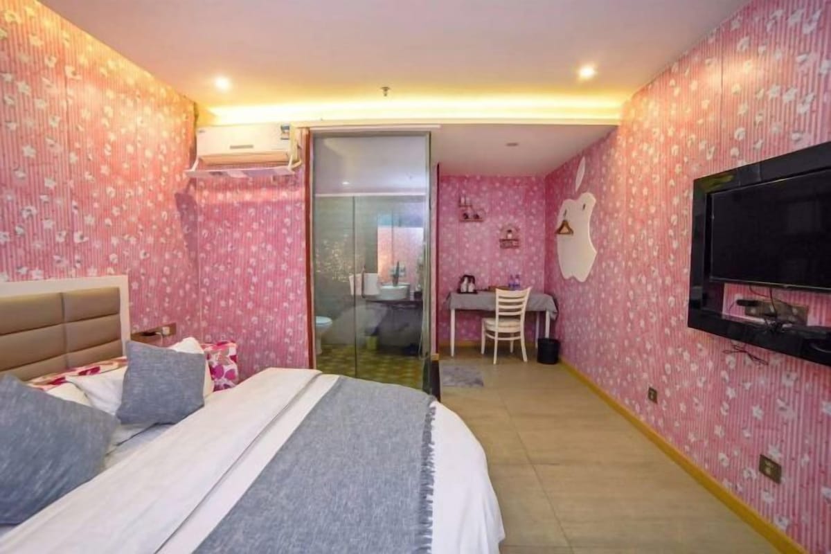 A bedroom with a bed, a desk, a chair, a television, and a bathroom visible through a glass door.