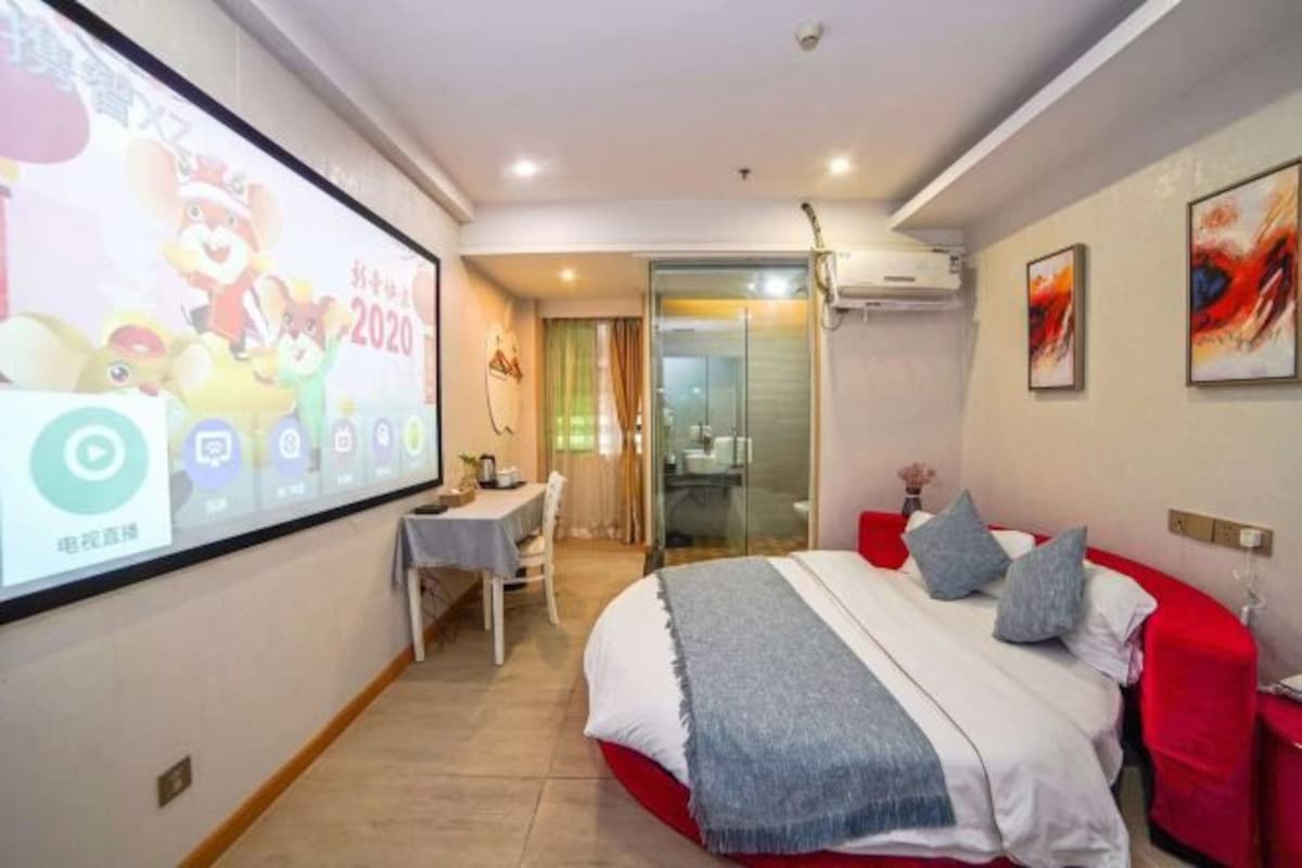 A hotel room with a bed, a desk, a chair, a TV screen, and a glass door leading to a bathroom.
