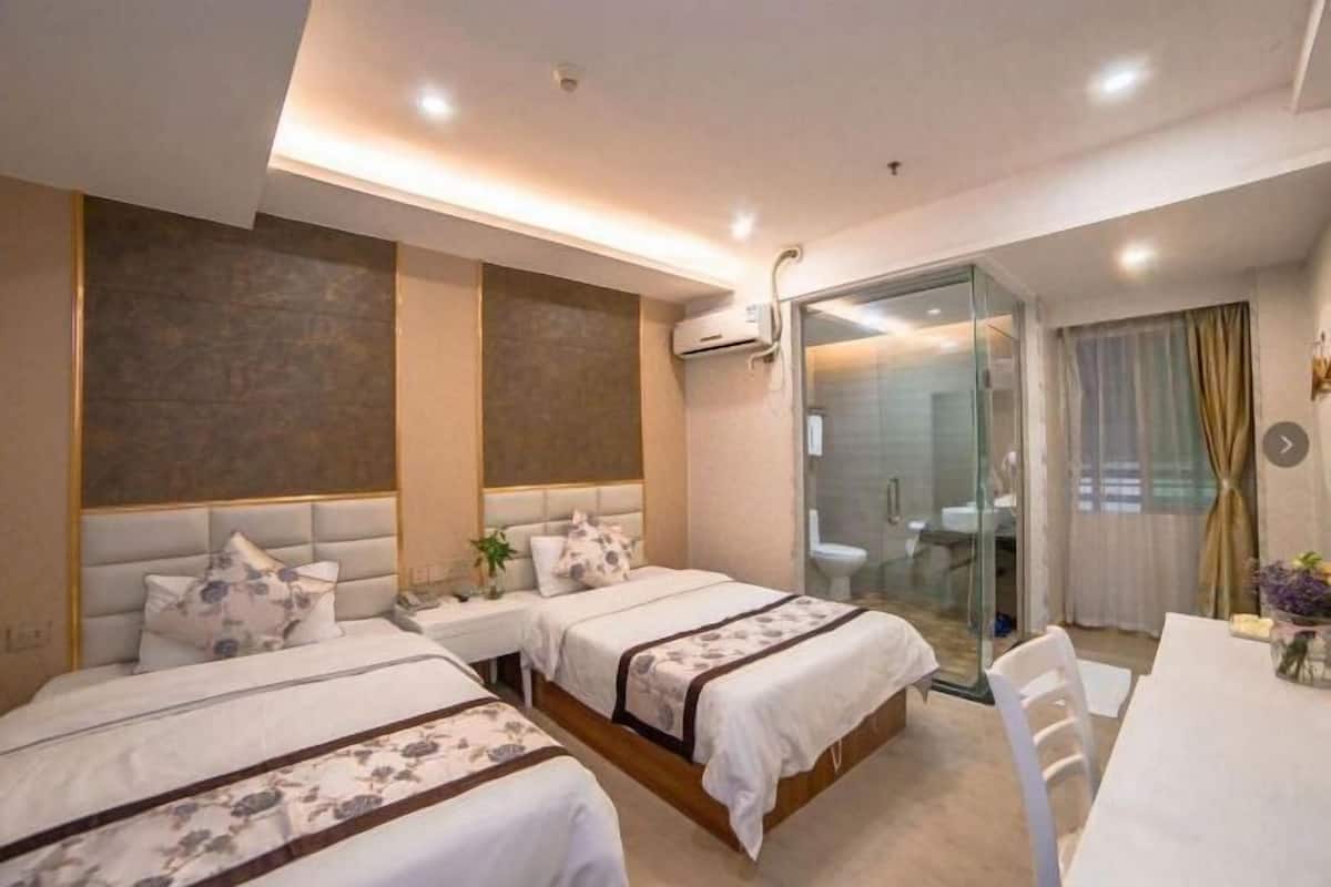 A hotel room with two beds, a desk, and a bathroom visible through a glass partition.