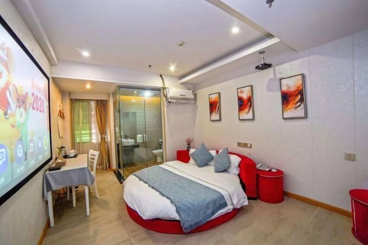 A modern bedroom with a circular bed, a glass shower, a dining area, and a TV screen displaying a presentation.