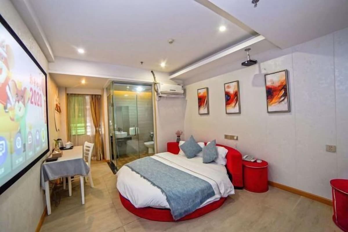 A modern bedroom with a circular bed, a glass shower, a dining area, and a TV screen displaying a presentation.