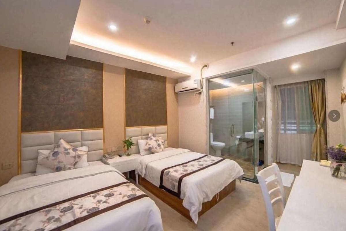 A hotel room with two beds, a desk, and a bathroom visible through a glass partition.
