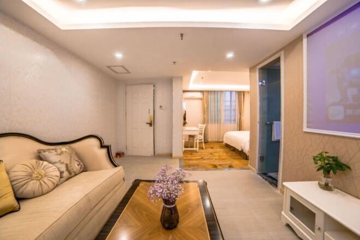 A hotel room with a sofa, a coffee table with a vase, a TV, a bed, and a bathroom visible through an open door.