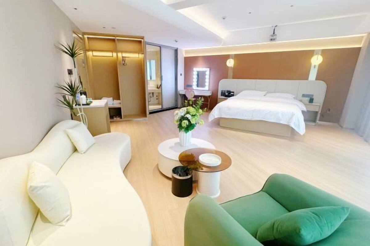 Une chambre d’hôtel moderne avec un lit, un canapé vert, un canapé blanc, une petite table ronde et un miroir.