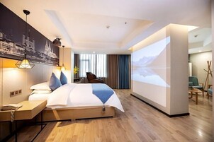 A modern hotel room with a large bed, a desk, a wall-mounted TV, and a cityscape painting.