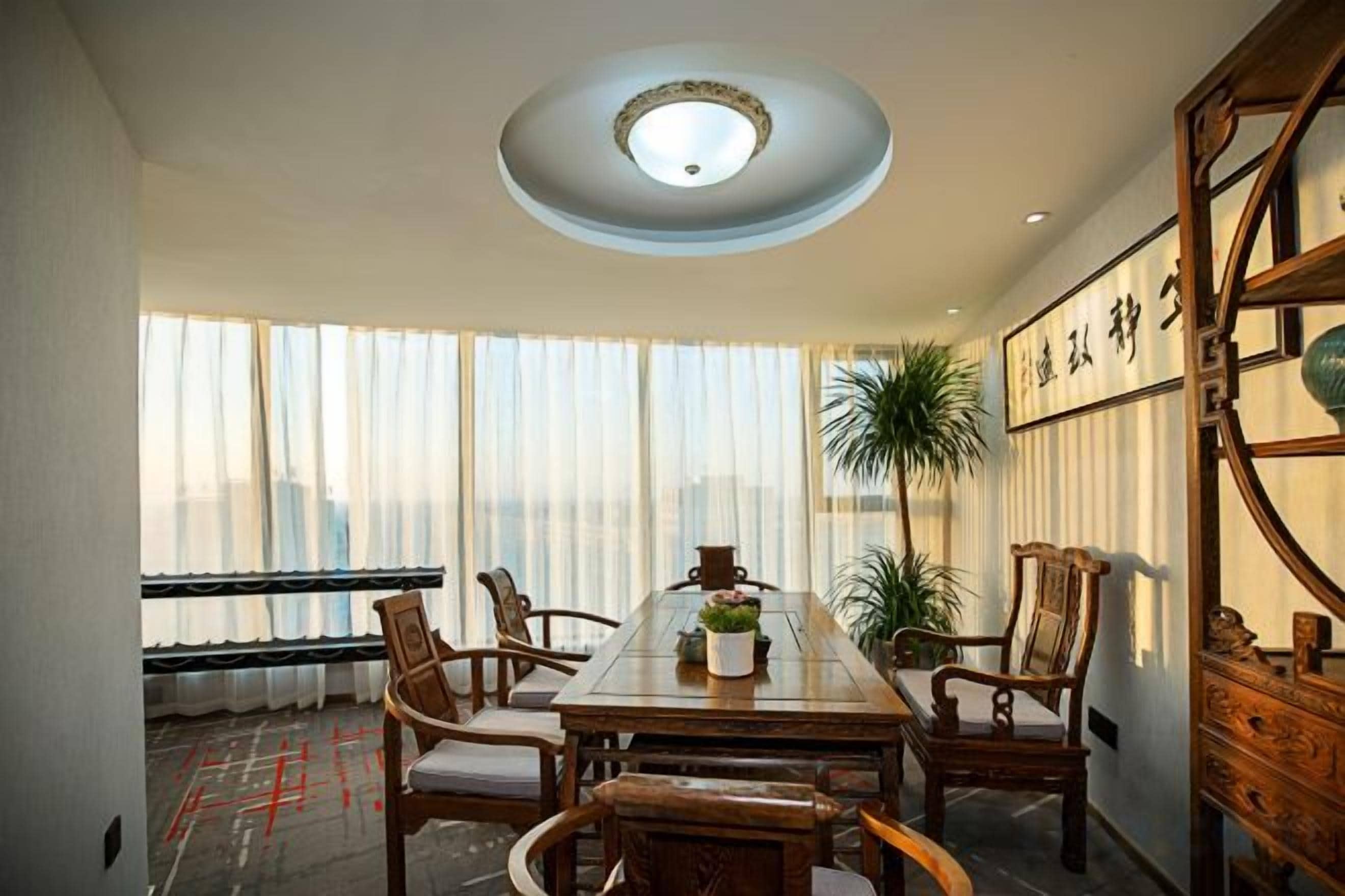 A dining area with a long table, chairs, a ceiling light, and a view through large windows.