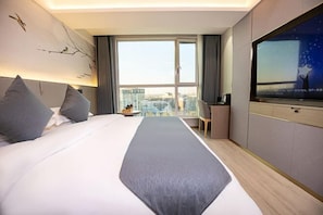 A hotel room with a large bed, a desk, a chair, a TV, and a view of the city through a large window.