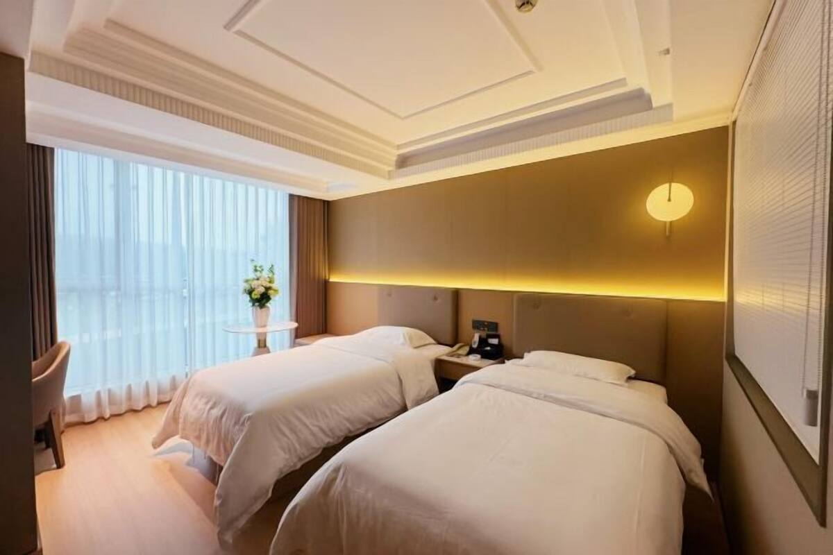 Une chambre d’hôtel avec deux lits, une petite table ornée d’un vase de fleurs, une table de chevet avec une lampe et une grande fenêtre avec des rideaux.