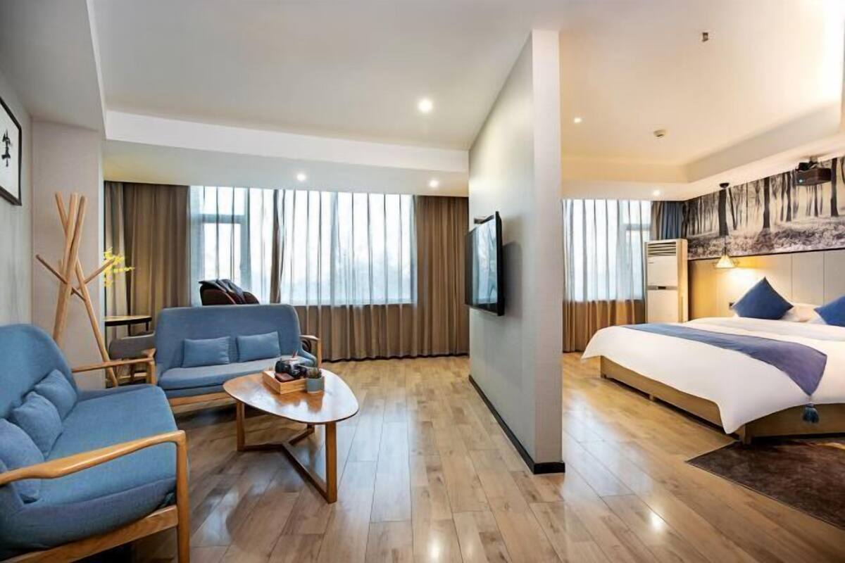 Une chambre d’hôtel moderne dotée d’un grand lit, d’un coin salon avec des canapés, d’une télévision à écran plat et d’une grande fenêtre avec des rideaux.