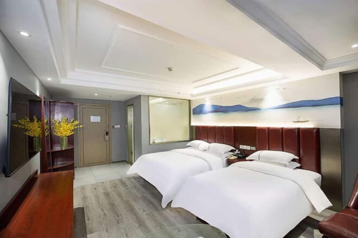 Une chambre d’hôtel avec deux lits, un banc en bois, un bureau, un miroir et un téléviseur fixé au mur.