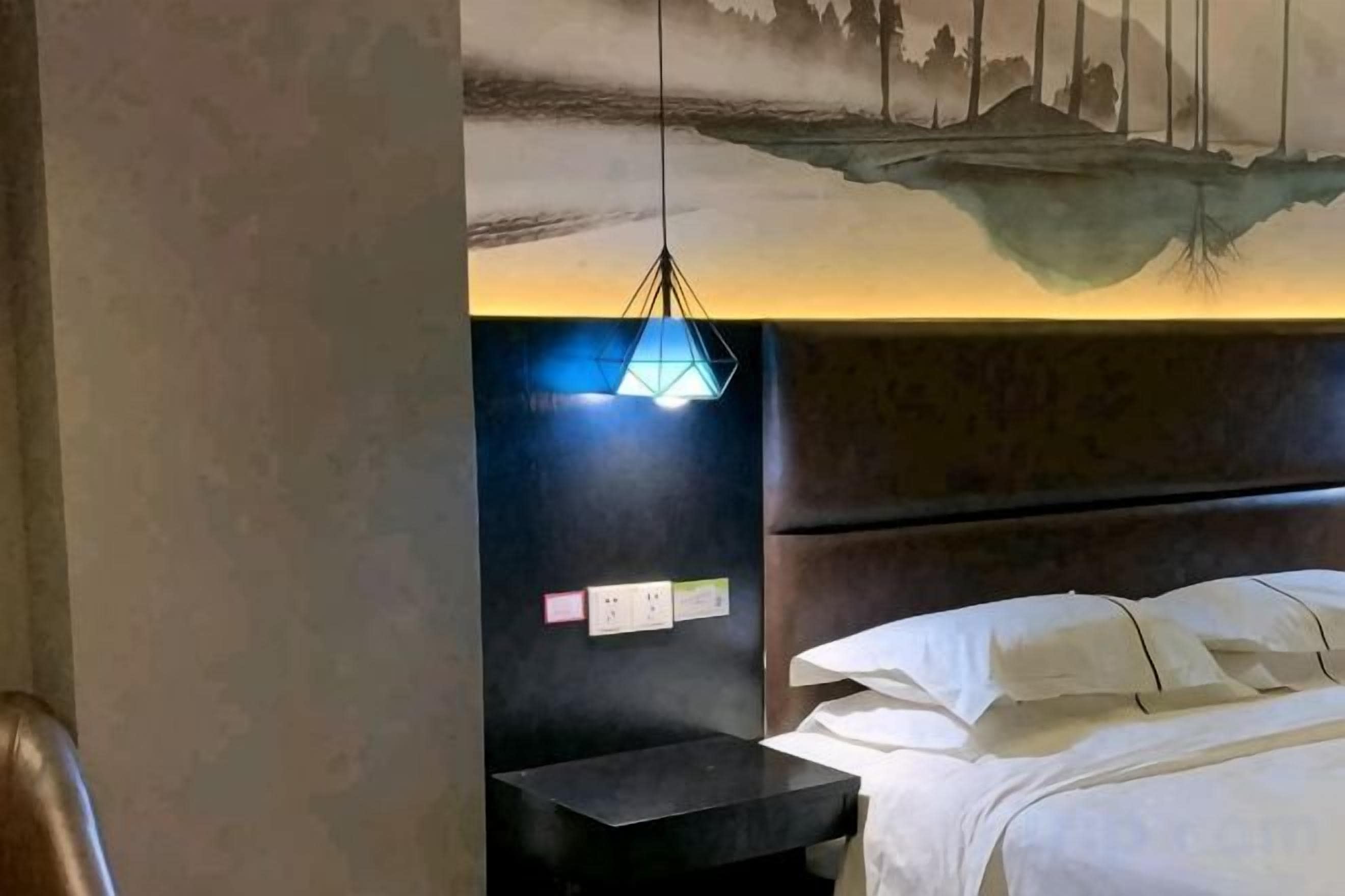 Quarto de hotel com cama, mesa de cabeceira, luminária na parede e um mural.