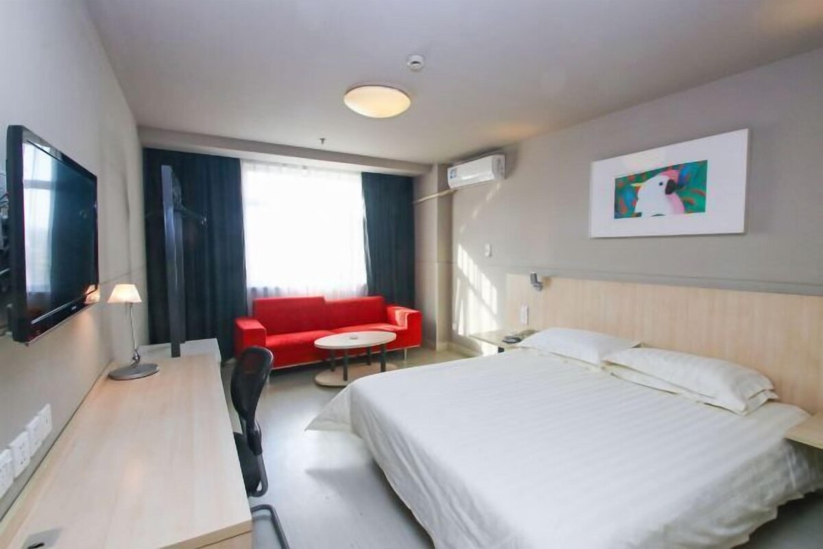 Quarto de hotel com cama, escrivaninha, sofá vermelho, televisão e um quadro na parede.