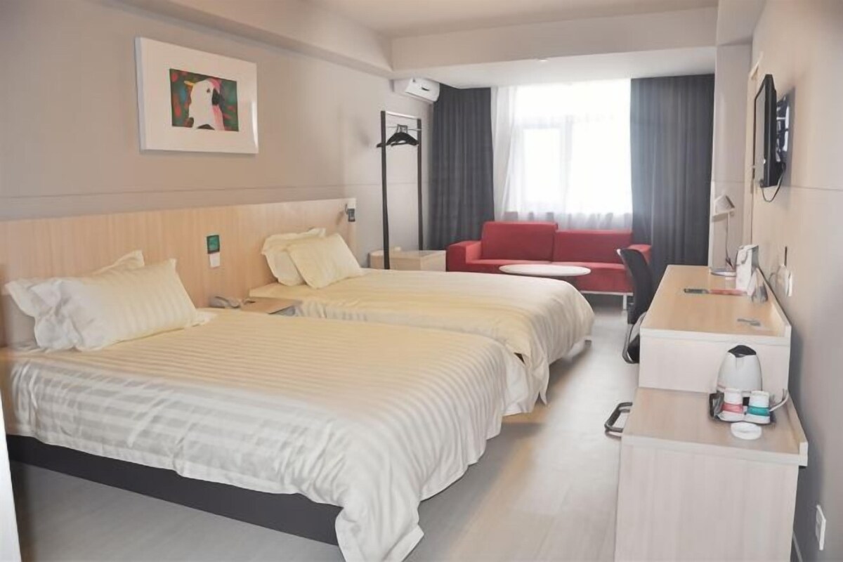 Une chambre d’hôtel avec deux lits, un bureau, une chaise, un canapé rouge et une fenêtre avec des rideaux.