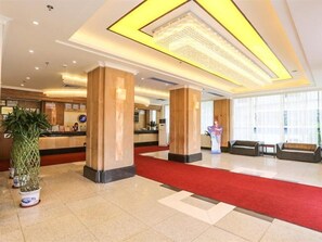 Una hall d'albergo con una reception, ampie colonne, un tappeto rosso e un'ampia finestra che offre luce naturale.