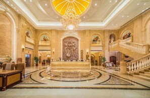 A grand lobby with a central reception desk, a large chandelier, and a staircase with balustrades.