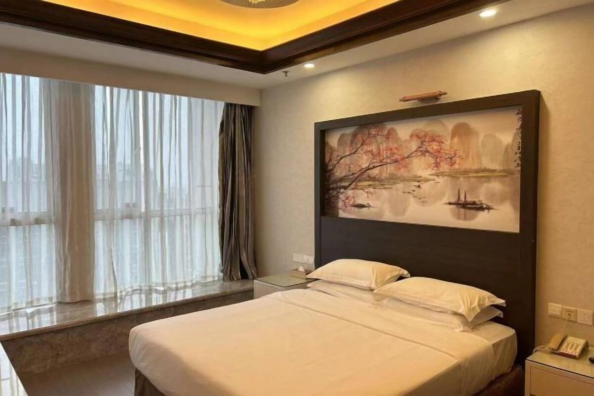 Una habitación de hotel con cama, un cuadro en la pared, una ventana con cortinas y una mesita de noche.