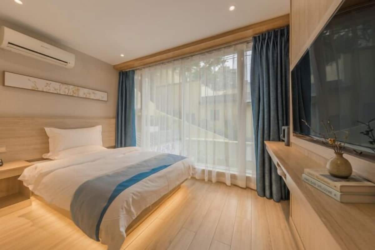 A modern hotel room with a large bed, a bedside table, a window with sheer curtains, and a wall-mounted air conditioner.