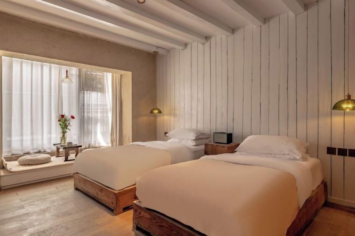 Una habitación con dos camas, una ventana con cortinas, una mesita de noche con una lámpara y una pequeña mesa con un florero.