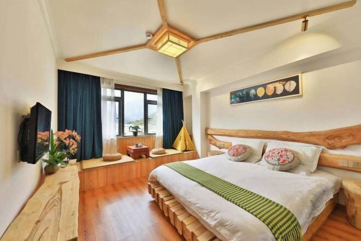 Ein Schlafzimmer mit Holzkopfstütze, einem Bett mit grüner Streifenoptik, einer Holzbank, einem Fenster mit Vorhängen und einem an der Wand hängenden Bild.