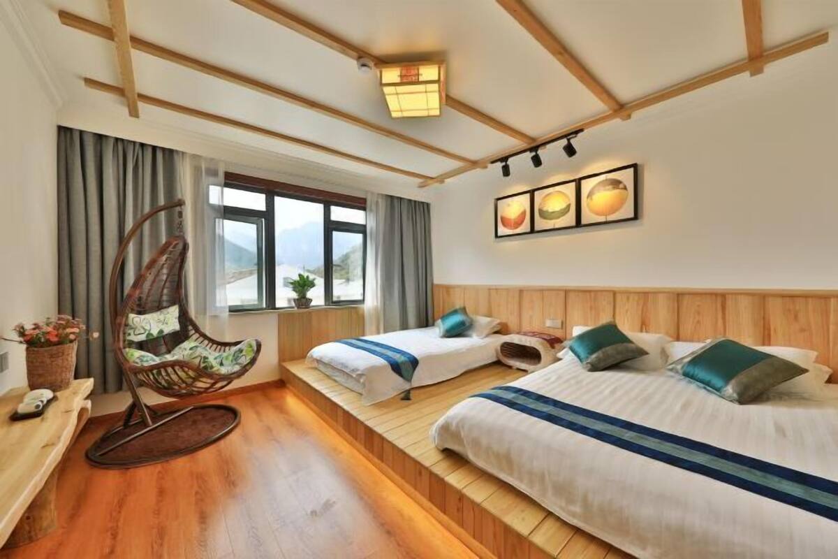 Ein Hotelzimmer mit Holzboden, zwei Betten mit blaugrünen Kissen, einem Schaukelstuhl, einem Fenster mit Vorhängen und einer Wandleuchte.