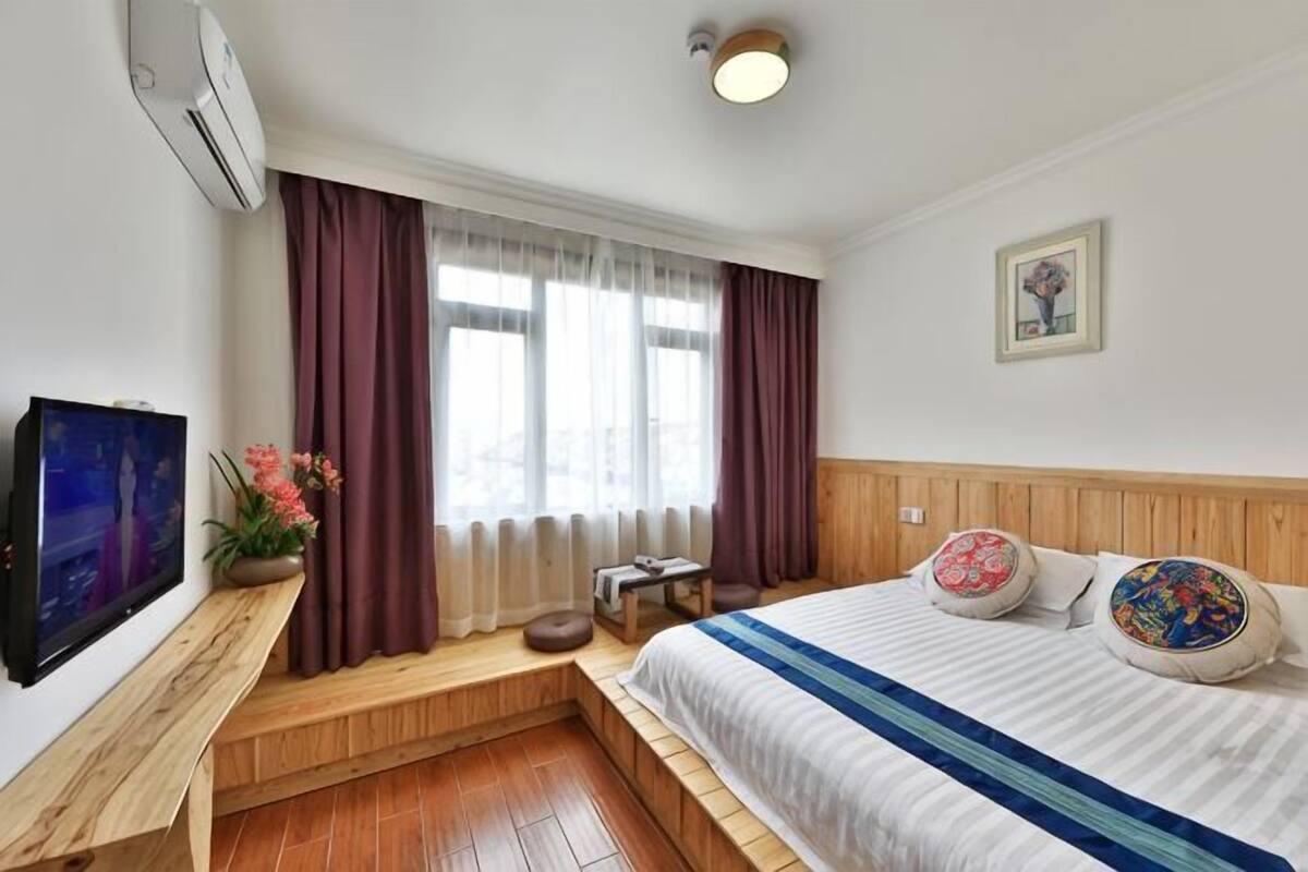 Ein Hotelzimmer mit Bett, Fernseher, Fenster mit Vorhängen, Nachttisch und einem Wandbild.