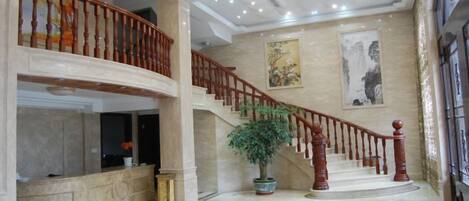 A grand lobby with a marble floor, a staircase with wooden railings, and a large potted plant.