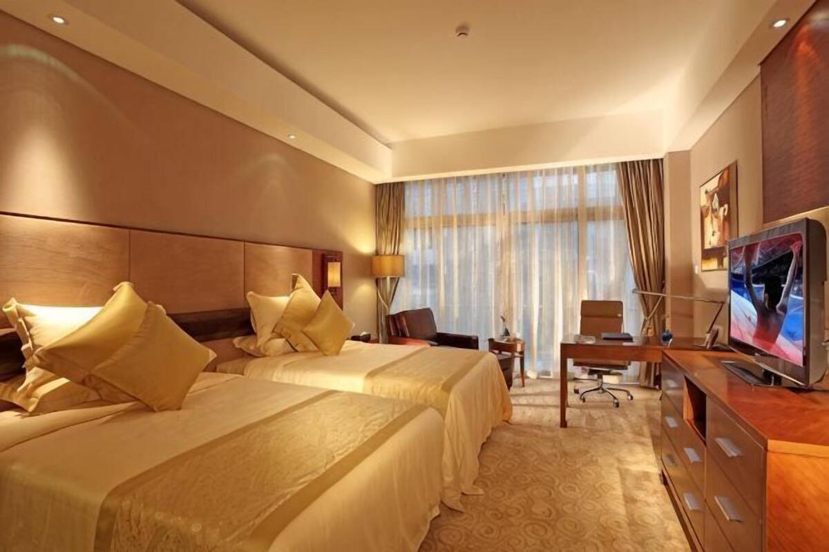 Una camera d'albergo con due letti, una scrivania, una TV e un'ampia finestra con tende.