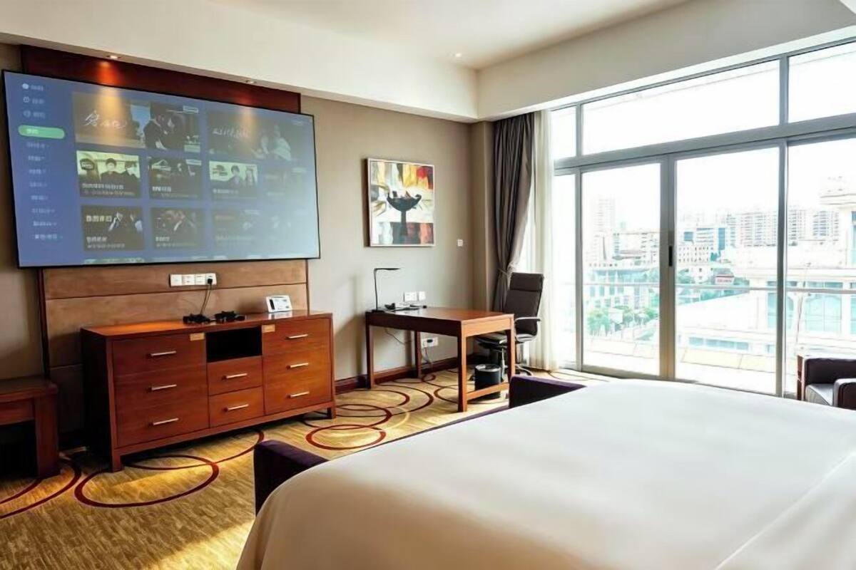 Camera d'albergo con un letto grande, una TV fissata al muro, una scrivania e vista sulla città attraverso ampie finestre.