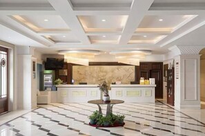 A hotel lobby with a reception desk, a marble counter, a round table with plants, and a tiled floor.
