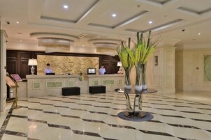 Zona de recepción de un hotel con mostrador, dos monitores, un jarrón grande con plantas y un piso con diseño.