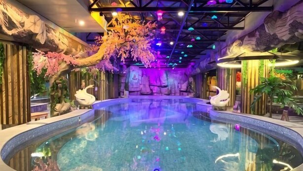 An indoor swimming pool with decorative swan statues, a ceiling with colorful lights, and a wall with a mural. - Royal Hot Spring Hotel (Baishan)