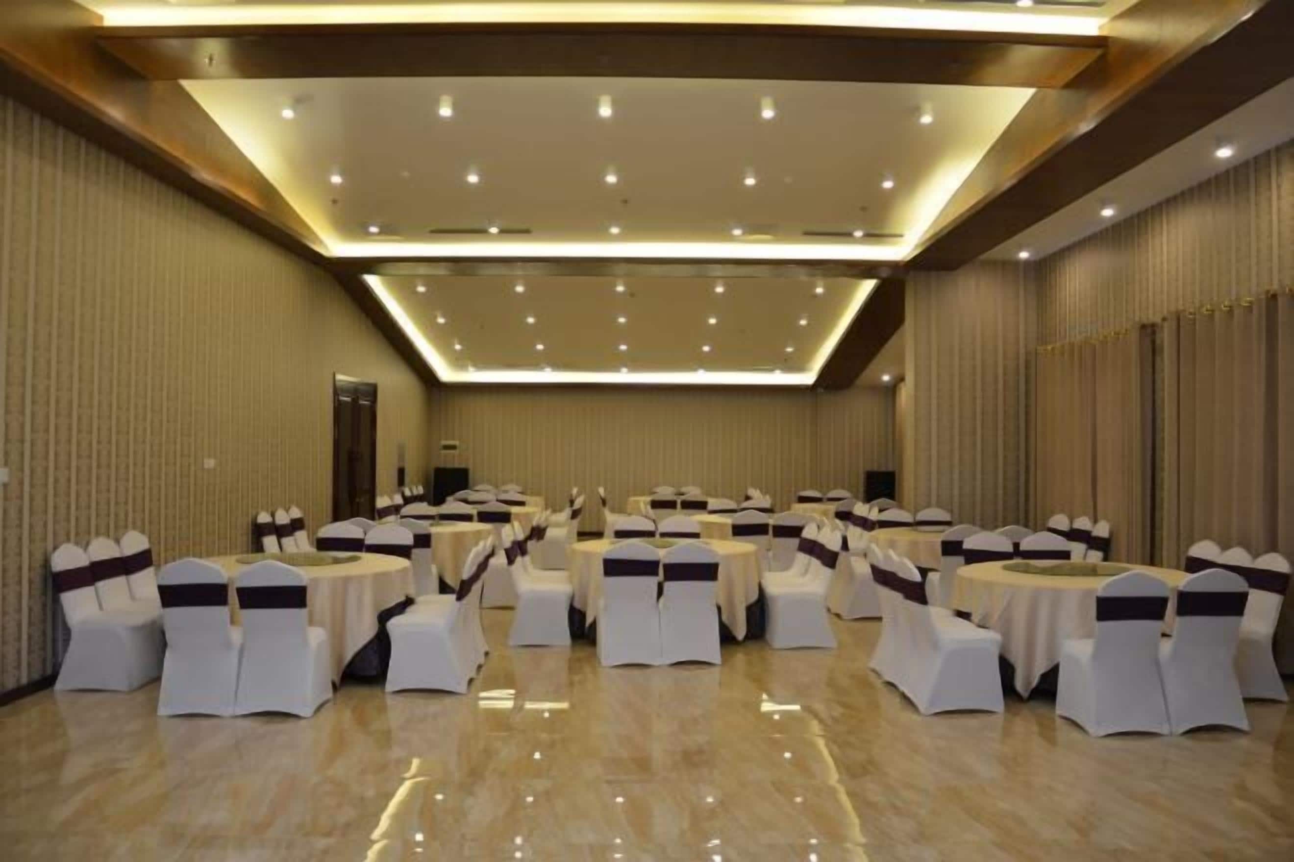 A large conference room with round tables and white chairs, each with a dark sash. The room has a high ceiling with recessed lighting and a polished floor.