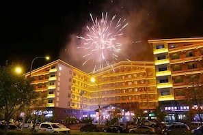 Um hotel com vários andares e entrada imponente, iluminado por postes de luz, com um grande espetáculo de fogos de artifício ao fundo.