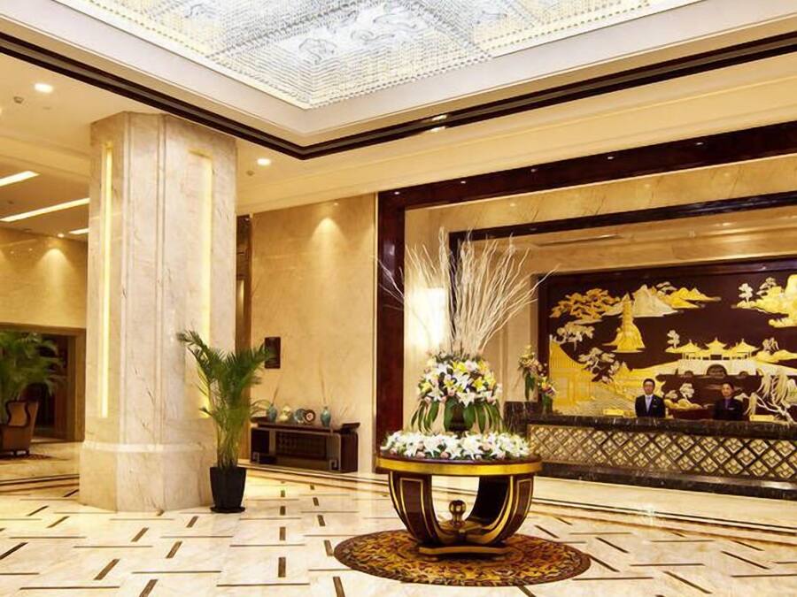 Un vestíbulo amplio con techo alto, una lámpara de araña grande, un muro decorativo con un mural dorado y una mesa redonda central con flores.