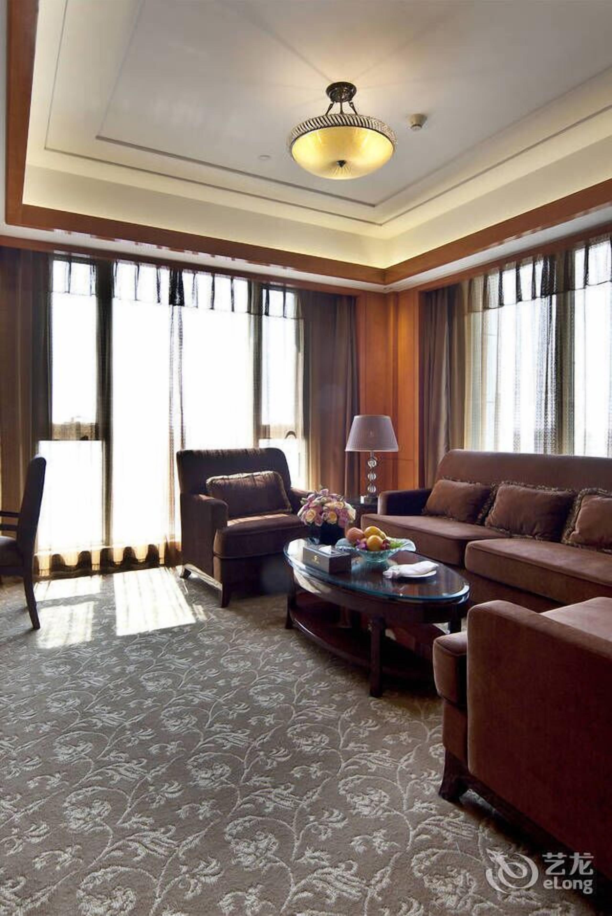 A hotel room with a large window, a sofa, a chair, a table with a lamp, and a patterned carpet.