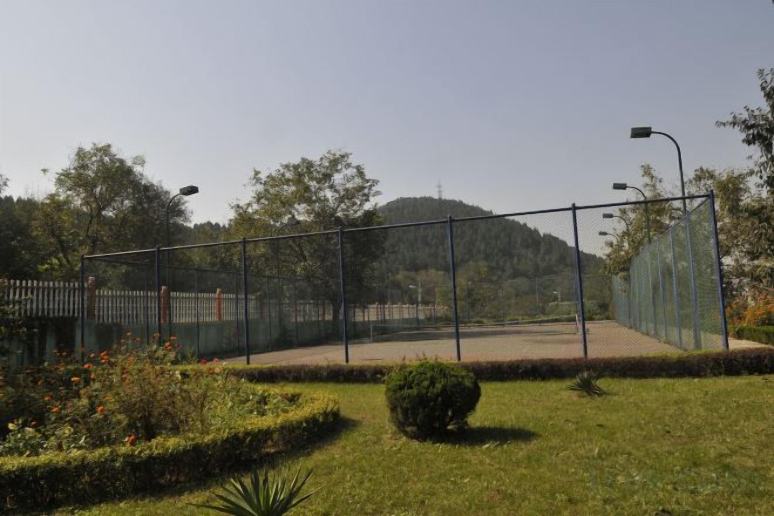 Una cancha de tenis cercada, con cielo despejado y árboles al fondo.