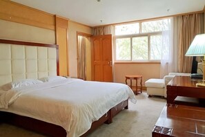 Quarto de hotel com uma cama grande, escrivaninha, cadeira, mesa pequena e janela com cortinas.