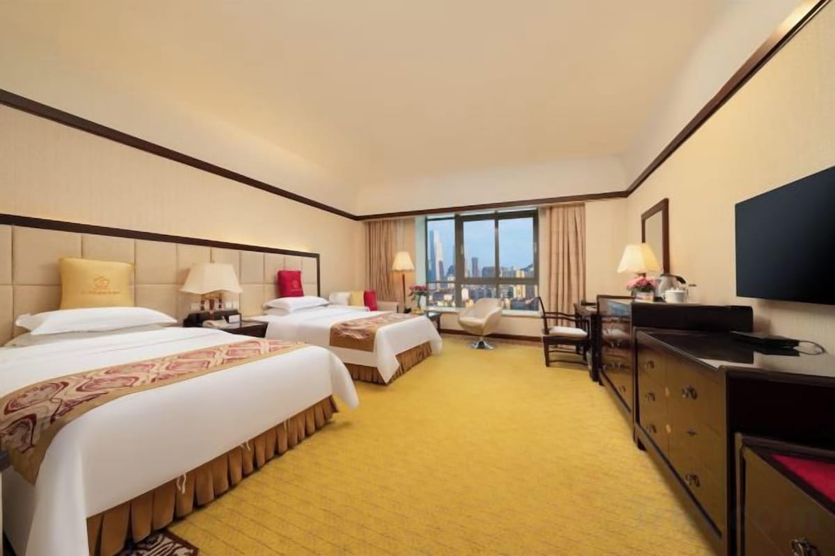 A hotel room with three beds, a TV, a desk, and a view of the city.